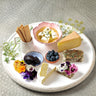 On a white large plat is laid Crackers, Persian Feta in a pink ramekin, Mature Cheddar, Various Chèvre, Monet with purple and pink flowers, Camembert sliced in half, star anise jam blue ramekin