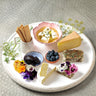 On a white large plat is laid Crackers, Persian Feta in a pink ramekin, Mature Cheddar, Various Chèvre, Monet with purple and pink flowers, Camembert sliced in half, star anise jam blue ramekin